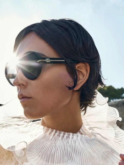 A woman with dark hair and fair skin wears sunglasses and a white ruffled top in an outdoor setting with a bright sunny sky.