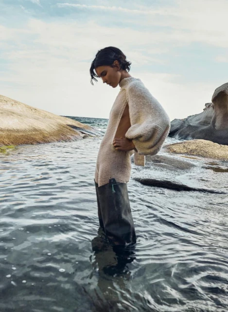 Woman in oversized knit sweater stands in shallow water near rocks under cloudy sky in a fashion or editorial shot.