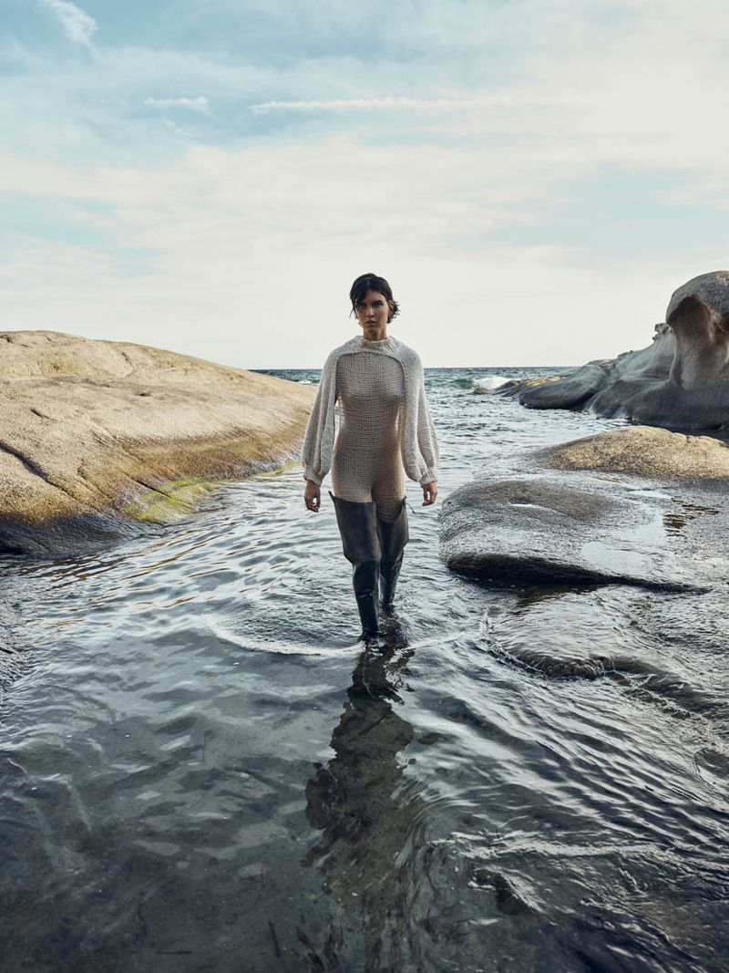 Model wading through shallow water between rocky coastal shores under a bright sky for Harper's Bazaar Arabia