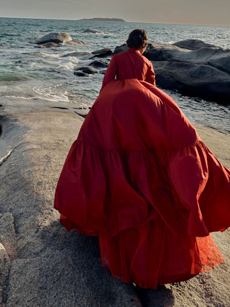 Woman in red gown standing on rocky shore looking out to sea in outdoor setting with natural light and island on the horizon.