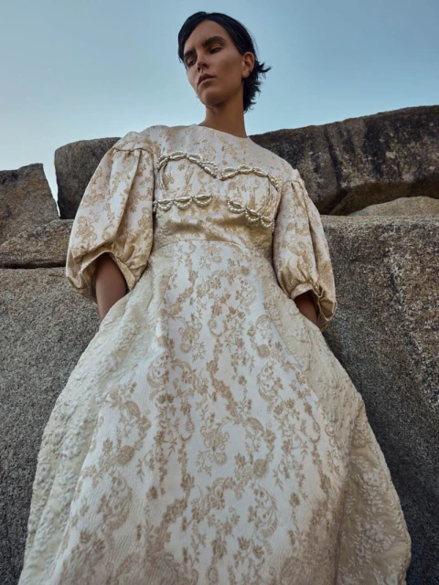 Young woman in an elegant beige dress stands near a weathered rock formation under a pale sky, creating a blend of fashion and nature.