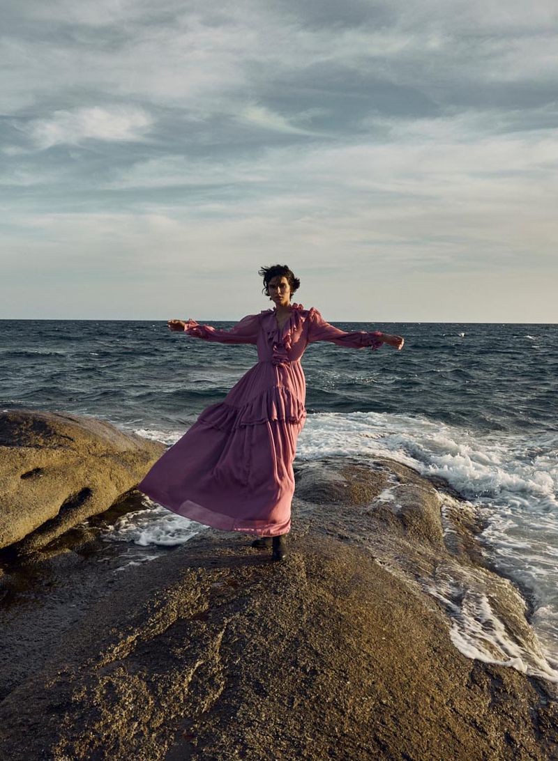 Model Polina Park posing on coastal rocks in a flowing mauve ruffled maxi dress against a seascape.