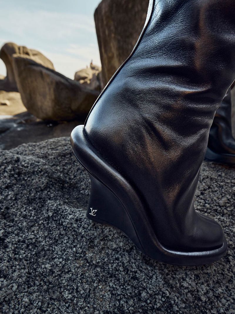 Close-up of a black leather wedge boot with a logo detail, set on rough terrain with blurred rock formations against a bright sky.