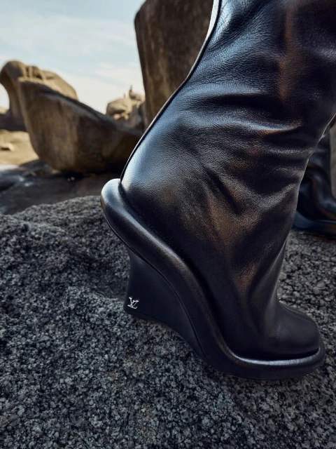 Close-up of a black leather wedge boot with a logo detail, set on rough terrain with blurred rock formations against a bright sky.