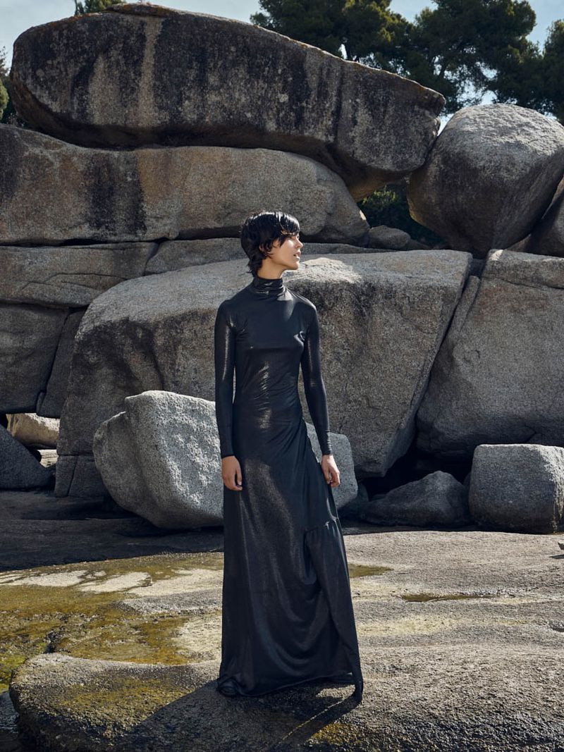 A full-length portrait of a young woman wearing a black dress, standing outdoors among large rock formations with trees in the background.