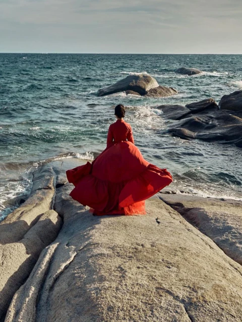 Woman in a red dress stands on a rocky shore, facing the ocean. The water is a deep blue with visible rocks in and near the water.