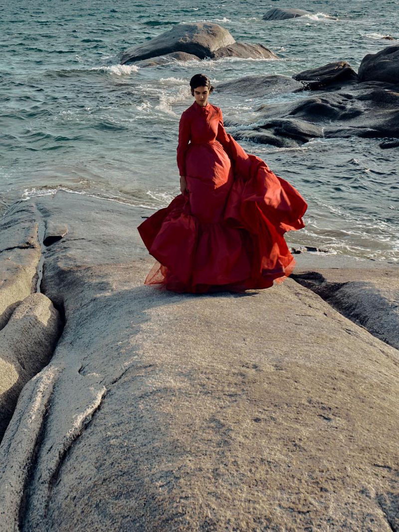 Woman in a red gown poses on a rocky outcrop by the ocean, waves visible in the background, creating a striking fashion editorial shot.
