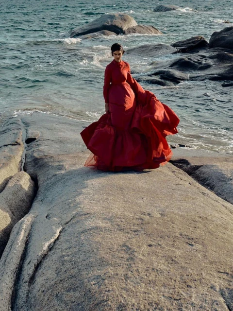 Woman in a red gown poses on a rocky outcrop by the ocean, waves visible in the background, creating a striking fashion editorial shot.