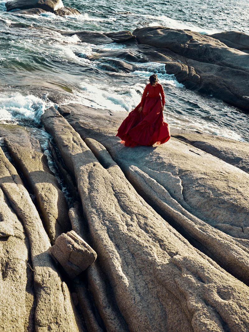 Model Polina Park wearing a flowing red gown walks across large coastal rock formations beside crashing ocean waves.