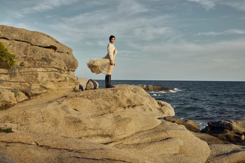 Woman in flowing dress stands on rocky coastline, gazing out at ocean under cloudy sky, wearing dark boots for fashion editorial.