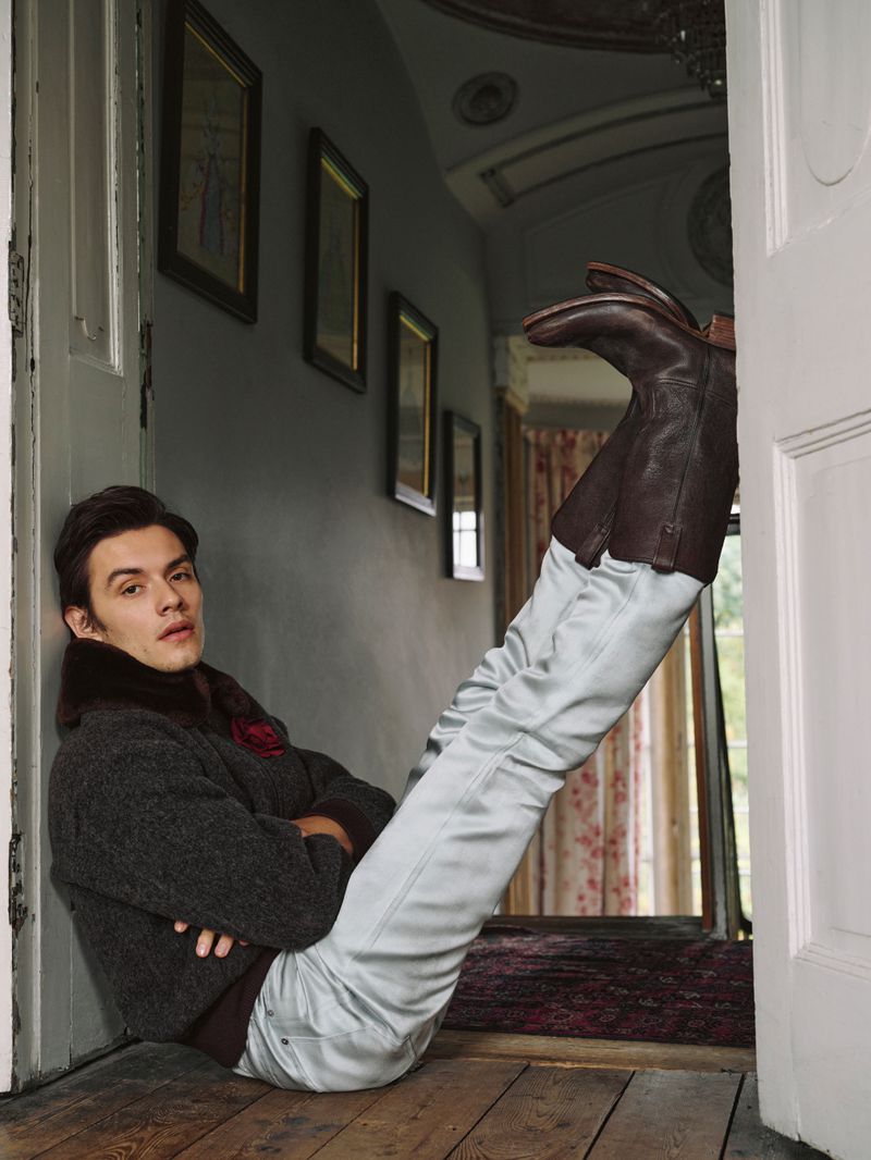 Stylish man in grey sweater and satin trousers poses indoors with brown leather boots.  Fashion editorial.  Grey walls, brown wood floor.