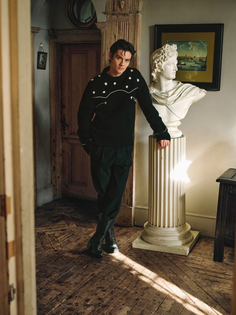 Indoor portrait of a young adult man leaning against a sculpture, wearing a black sweater and dark pants, standing on a wooden floor.