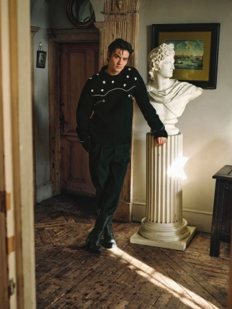 Indoor portrait of a young adult man leaning against a sculpture, wearing a black sweater and dark pants, standing on a wooden floor.