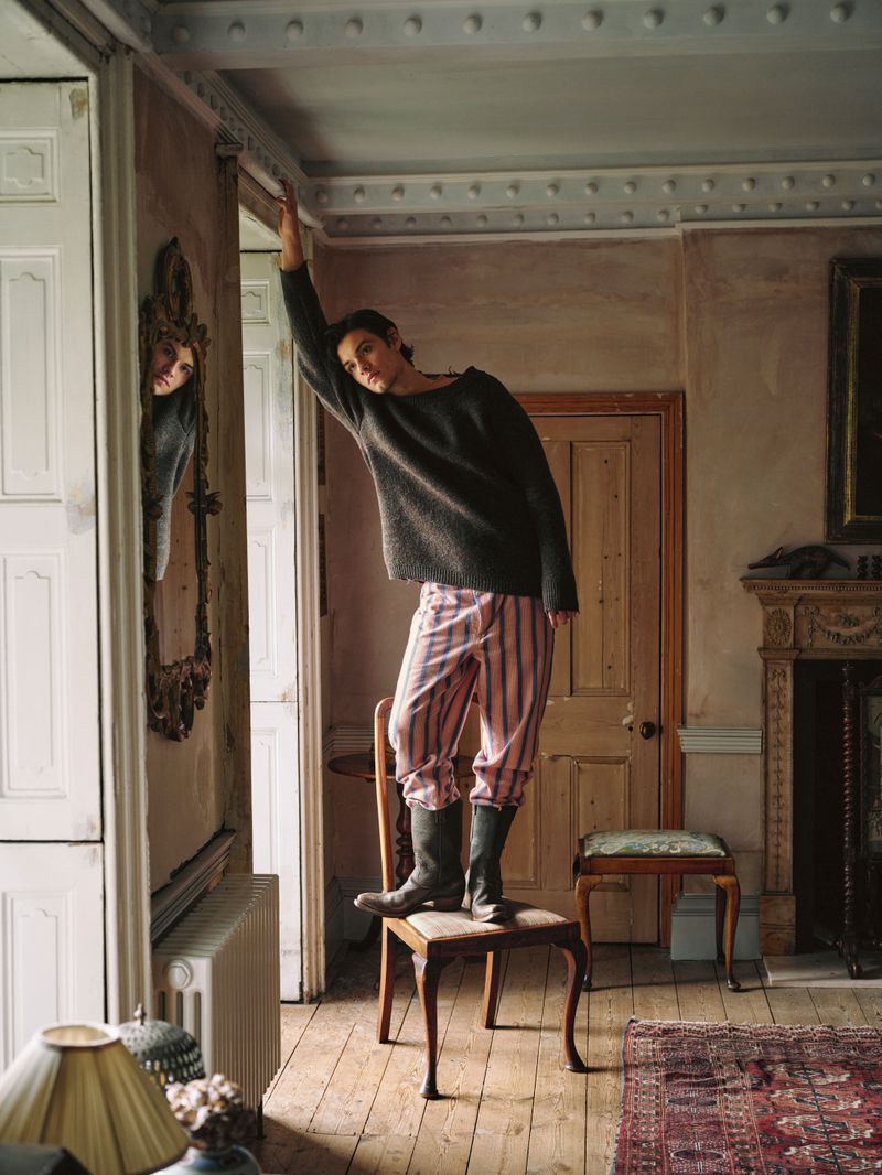Adult stands on chair in room with period details, wearing dark sweater, striped pants and dark boots. Mirror on wall reflects person.