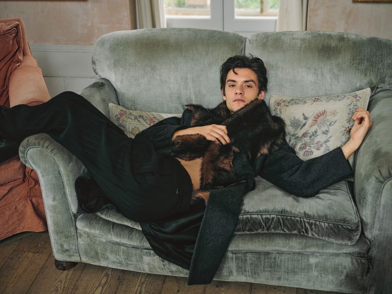 Brunette man in a gray coat and dark pants, lounging on a light-green velvet couch with a decorative pillow in a neutral-toned interior.
