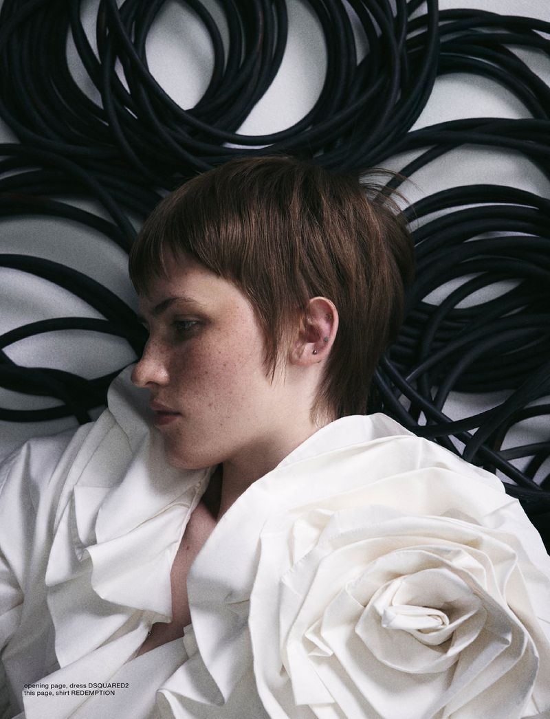 A young woman with short brown hair poses with a modern ruffled, rose-like white collar against a background of coiled black cables.