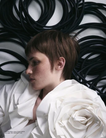 A young woman with short brown hair poses with a modern ruffled, rose-like white collar against a background of coiled black cables.