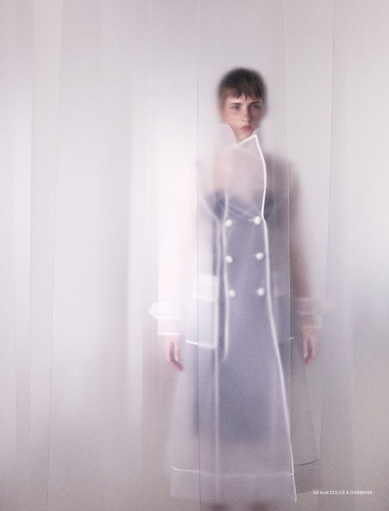 Woman in a translucent white coat stands in a white studio, creating a blurred, ethereal fashion portrait with diffused lighting.