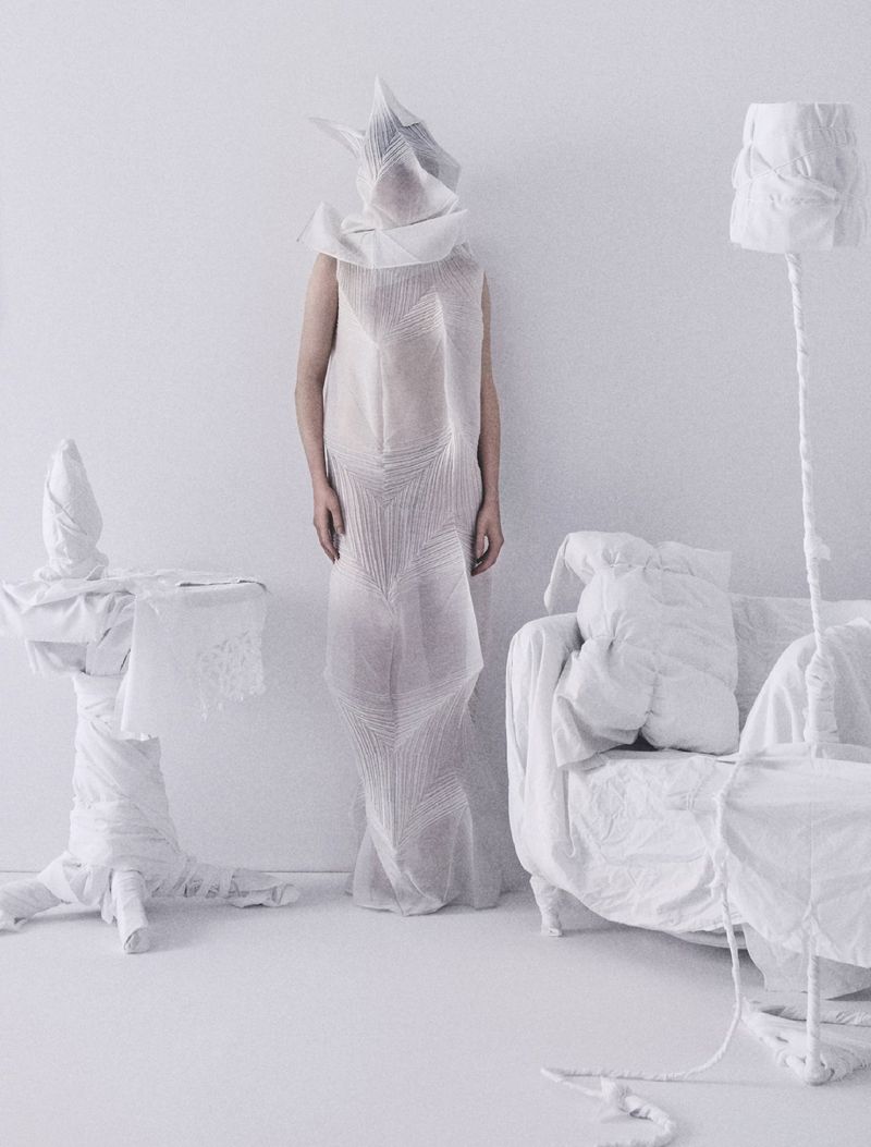 Woman wearing a draped white dress and head covering poses in a minimalist white space next to a draped couch, table, and lamp.