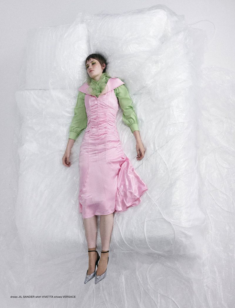 A woman in a pink dress and silver heels lies on a white, transparent surface in a studio, shot with a high angle.
