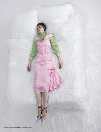 A woman in a pink dress and silver heels lies on a white, transparent surface in a studio, shot with a high angle.