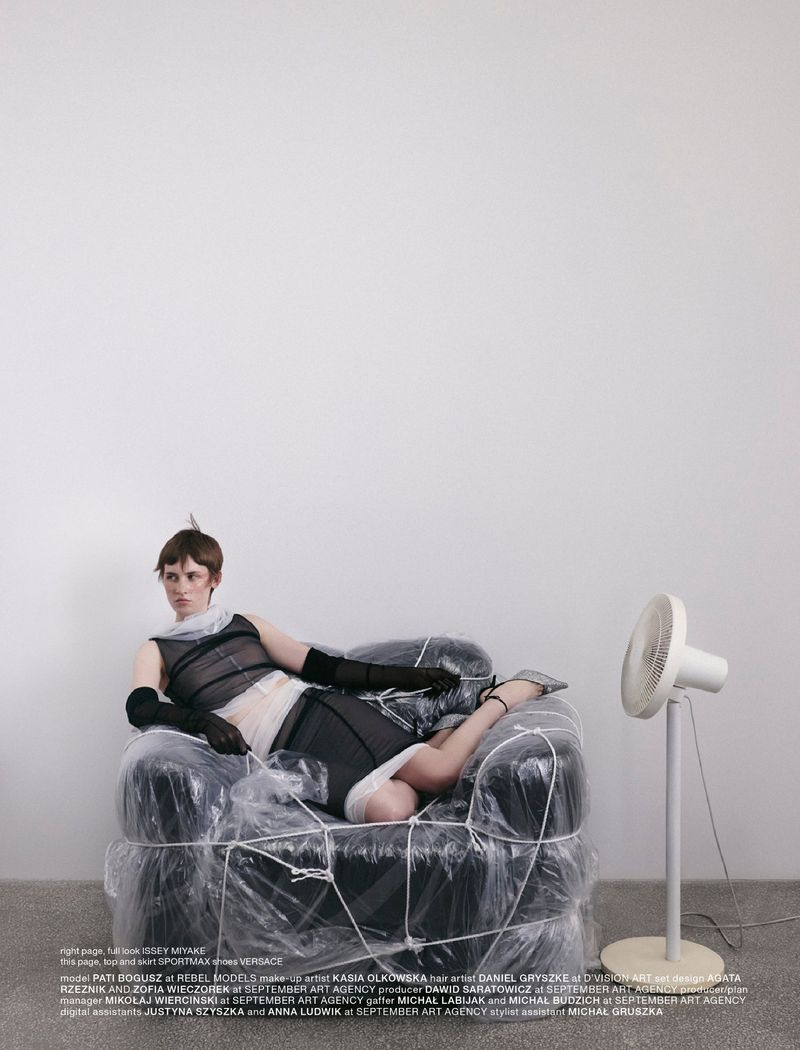Fashion editorial image featuring a woman in black and white outfit reclining on a chair wrapped in plastic, next to a white fan.