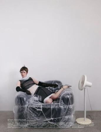 Fashion editorial image featuring a woman in black and white outfit reclining on a chair wrapped in plastic, next to a white fan.