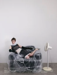 Model reclining in a plastic-wrapped armchair with a white standing fan against a plain wall for Numéro Switzerland.