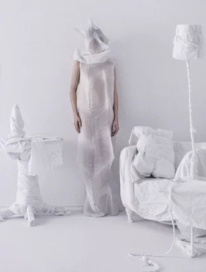 A figure in a textured sheer white gown and matching fabric headdress stands in an all-white room with fabric-covered furniture.