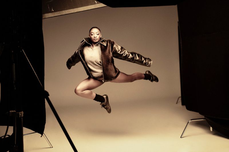 Athletic woman in a light brown bomber jacket and athletic shoes leaps with pointed toes in a studio with lighting equipment.