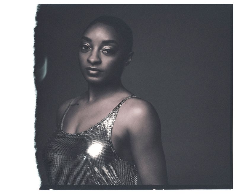 Portrait of Simone Biles wearing a metallic sequin top against a dark background in a square polaroid frame.