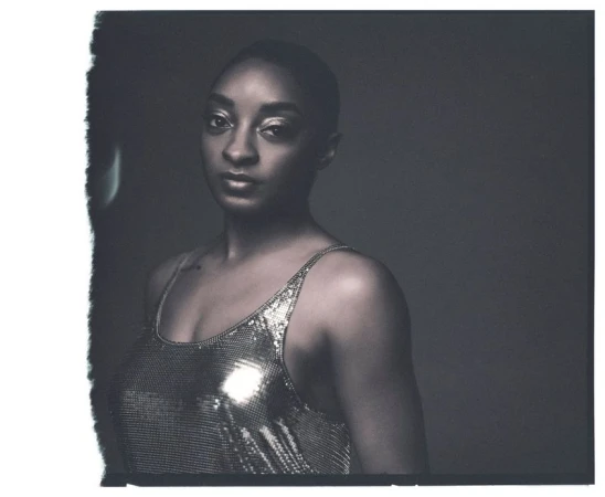 Portrait of Simone Biles wearing a metallic sequin top against a dark background in a square polaroid frame.