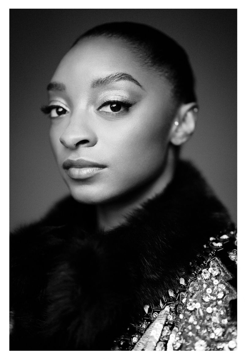 Close-up black and white portrait of a woman with dark hair pulled back, wearing a fur collar and embellished top against a grey backdrop.