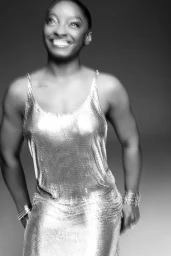 Black and white portrait of Simone Biles smiling in a shimmering metallic chainmail dress against a studio backdrop.