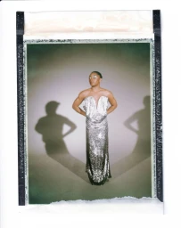 Simone Biles poses in a strapless silver sequin gown casting dual shadows on a studio backdrop in a Polaroid-style frame.
