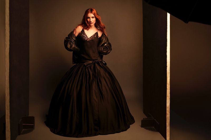 Red-haired woman in a black dress and leather jacket poses in a photo studio against a grey backdrop with studio lighting visible.