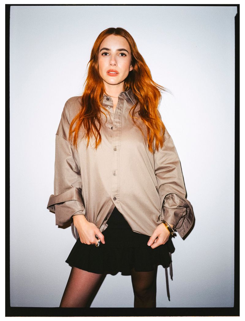 Portrait of a fair-skinned woman with red hair in a tan button-down shirt, black skirt, and tights, posing against a white backdrop.