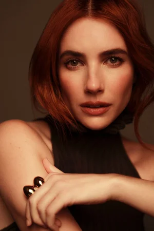 Emma Roberts with auburn red hair wearing a black sleeveless high-neck top and a gold ring in a studio portrait.