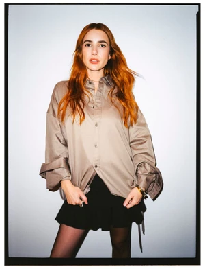 Emma Roberts wearing a beige button-down shirt and black skirt against a plain white studio wall