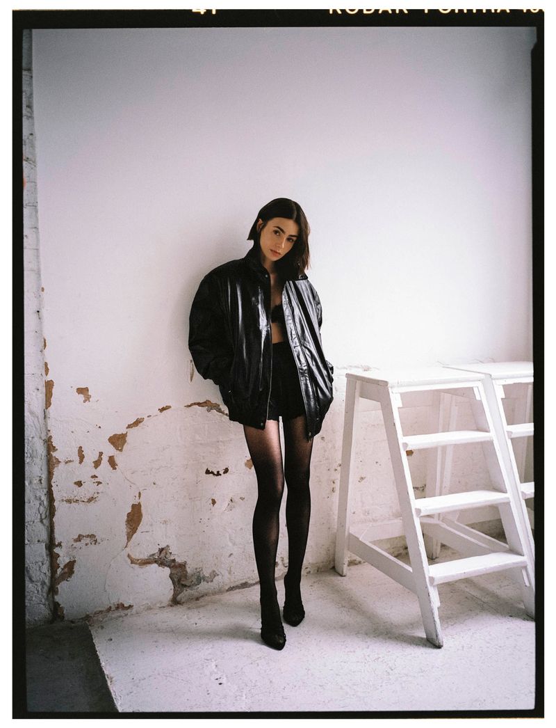 A young woman posing in a photo studio, wearing a black leather jacket, bra top, and shorts with sheer stockings in soft natural light.