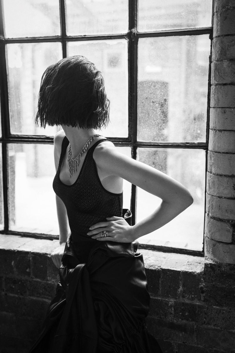 Black and white fashion portrait of an adult female in an elegant gown and statement necklace standing near a window, facing away from the camera.