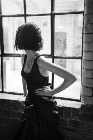Black and white fashion portrait of an adult female in an elegant gown and statement necklace standing near a window, facing away from the camera.