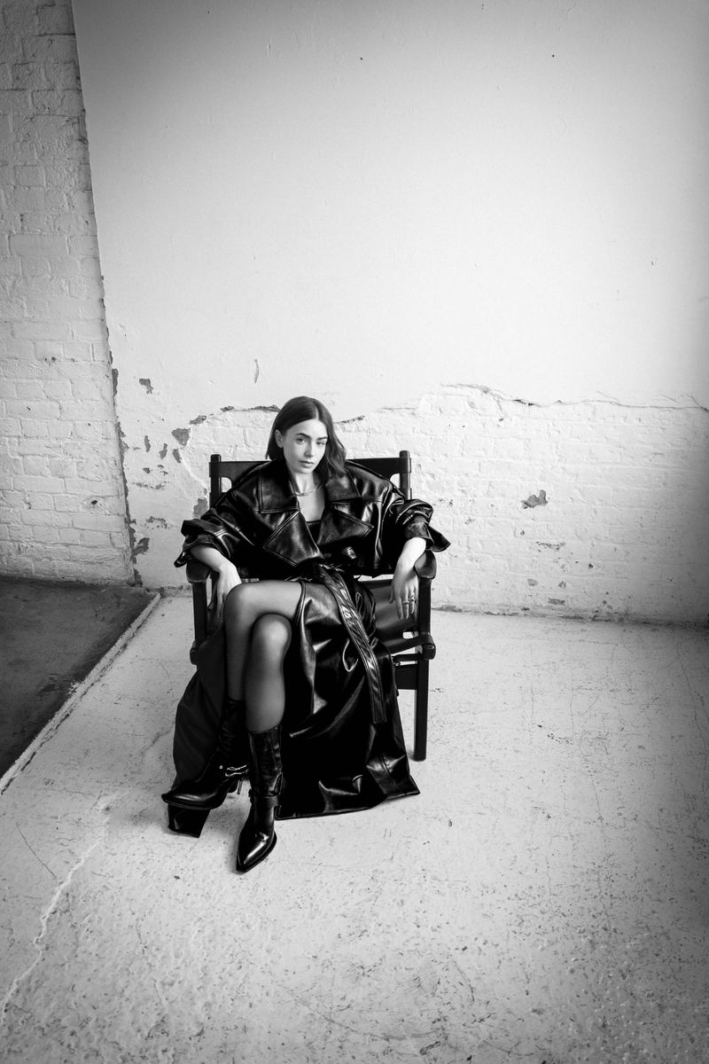 Monochrome portrait of a woman in a black coat sitting in a chair against a textured wall, shot in a studio with natural light.