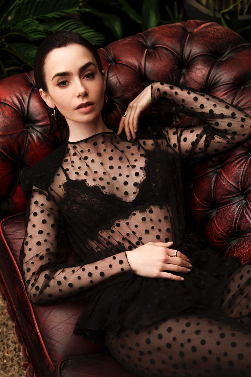 Lily Collins poses on a red leather couch wearing a black polka dot top. The background features green foliage, creating an elegant, lush scene.