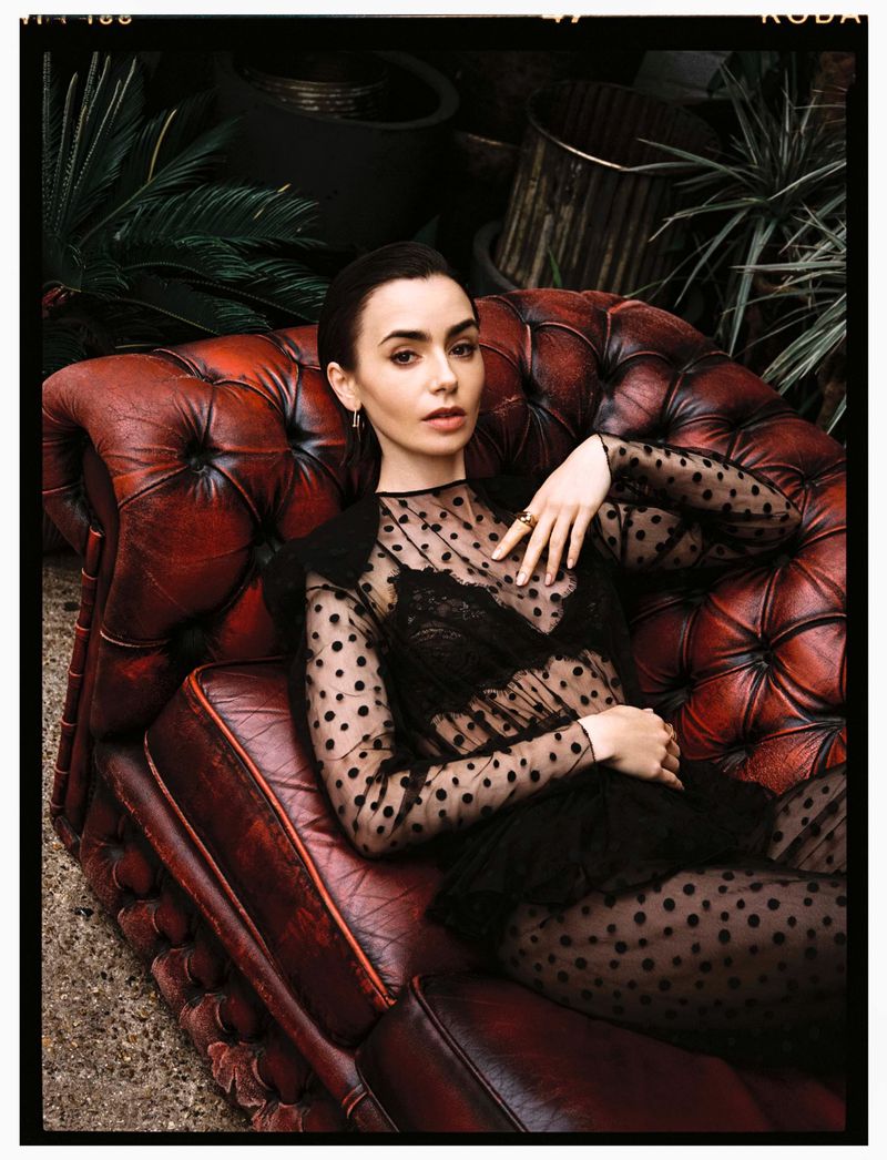 Elegant photo of a stylish woman with dark hair reclining on a red leather couch, wearing a black sheer polka dot dress in an indoor setting.