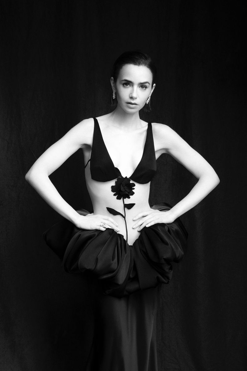 Monochrome portrait of a woman in a sculptural dress with flower detail, against a dark backdrop, emphasizing fashion and artistic expression.