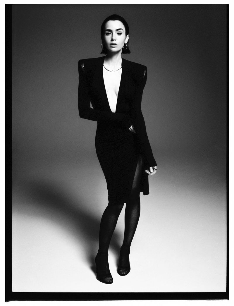 Portrait of a young woman in a black dress with a deep V-neck, accessorized with earrings and a necklace, posing in a photo studio.