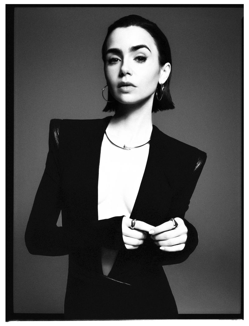 Elegant monochrome studio portrait of a woman with dark hair wearing a black blazer, gold earrings, and a delicate necklace, looking at the camera.