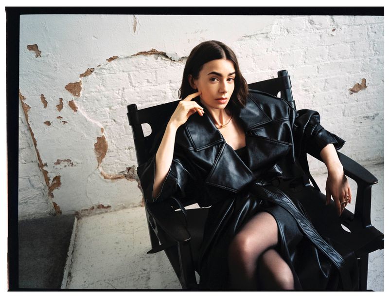 Lily Collins lounges in a black leather trench coat and dress in an arm chair against a distressed white wall in a fashion portrait.