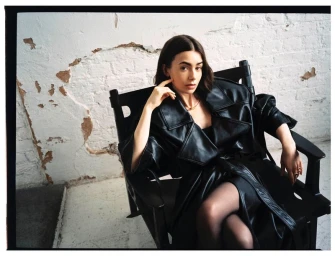 Lily Collins seated in a black chair wearing an oversized black leather trench coat against a distressed white brick wall.